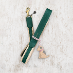 A green and beige leather dog leash with a gold-tone clasp, braided trim, and a hanging dog-shaped charm tied with pink-and-white string, photographed on a light rustic wood background.