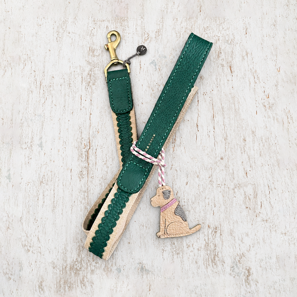 A green and beige leather dog leash with a gold-tone clasp, braided trim, and a hanging dog-shaped charm tied with pink-and-white string, photographed on a light rustic wood background.